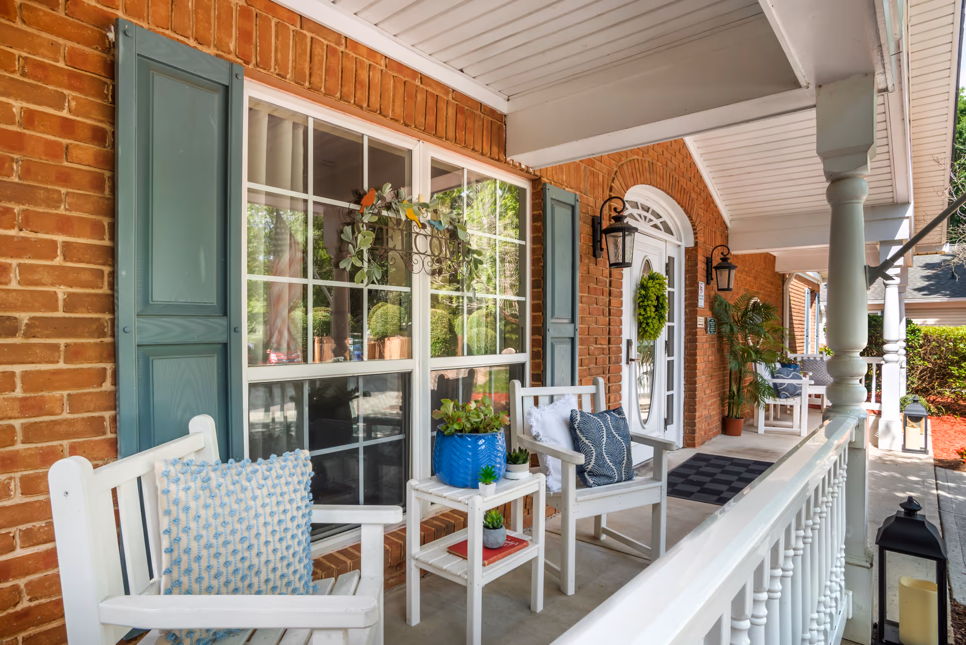 A cozy front porch with white wooden chairs adorned with decorative pillows, a small white table holding potted plants, and a brick exterior wall with teal shutters. The porch has white railings and columns, a white door with a green wreath, and black lantern-style wall lights. There are plants and greenery visible in the background.
