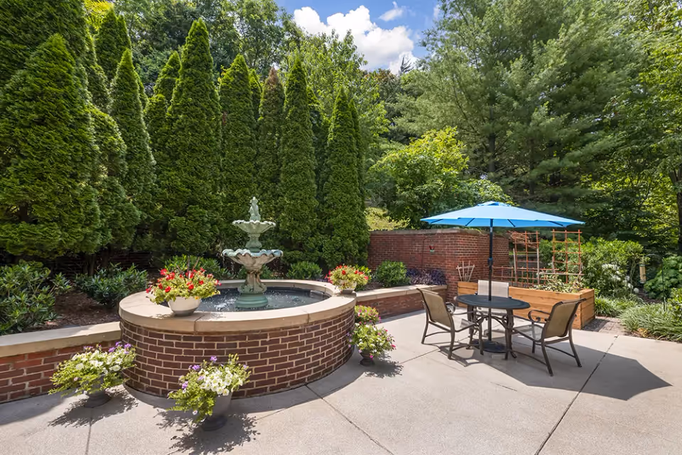 Outdoor patio area with a round brick water fountain surrounded by flower pots, a table with four chairs, and a blue umbrella. Tall green trees and shrubs provide a lush backdrop under a partly cloudy sky.