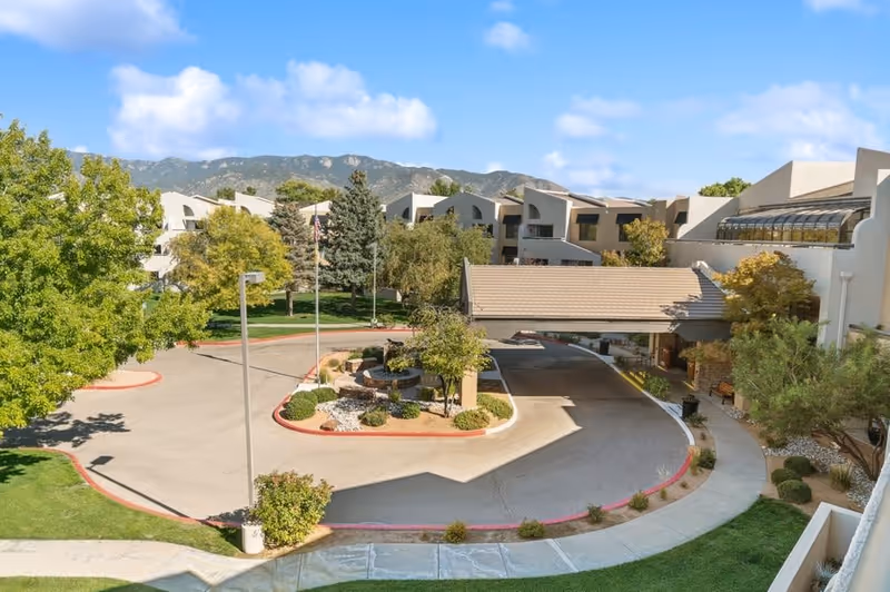 View of the exterior entrance area of The Montebello on Academy facility with a circular driveway, landscaped greenery, trees, and a covered drop-off area. Mountains and a partly cloudy sky are visible in the background.