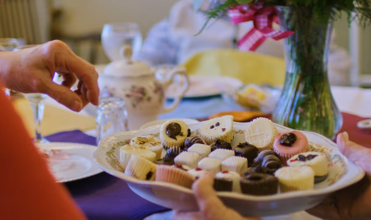A close-up of a plate filled with assorted small desserts and chocolates being held by a person, with another person reaching to take one. In the background, there is a table set with a teapot, glasses, plates, and a vase with flowers tied with a pink ribbon.