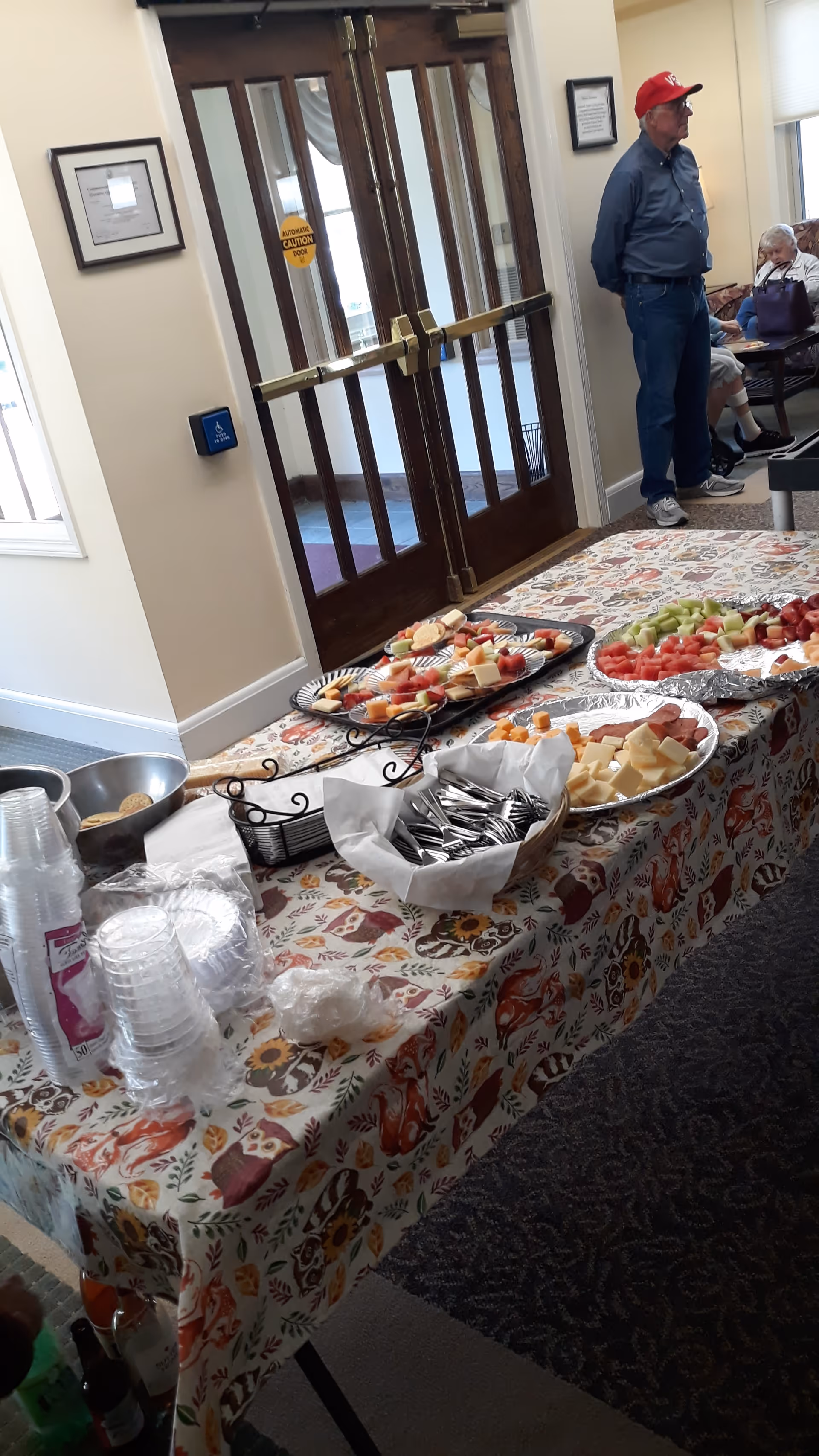 A table covered with a patterned tablecloth displaying trays of assorted fruits, cheeses, and crackers. Clear plastic cups, plates, and utensils are arranged on the table. In the background, there is a wooden double door with glass panels and a man wearing a red cap standing near a seated elderly person in a common area.