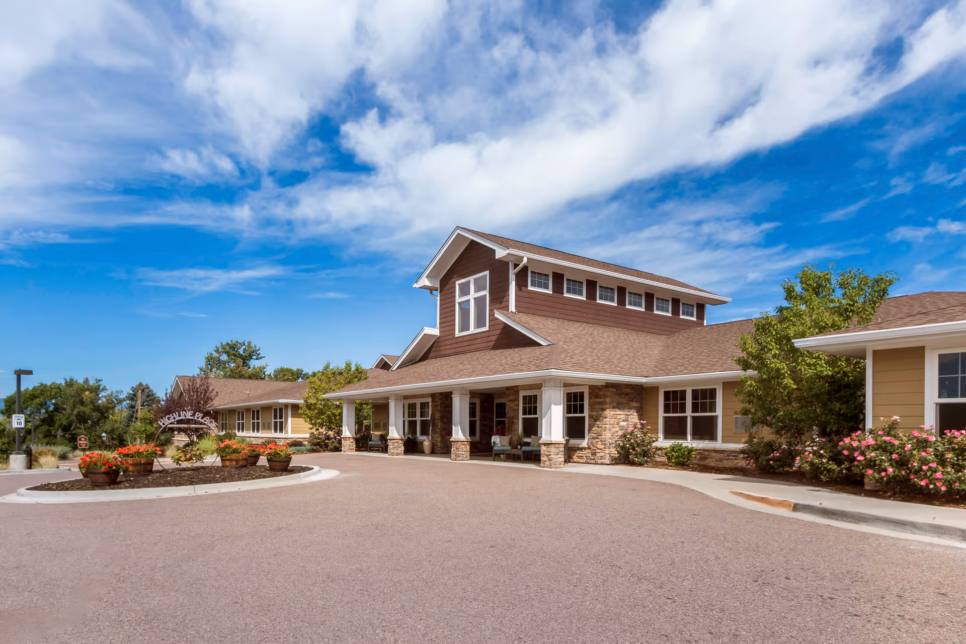 Exterior view of Highline Place Memory Care facility showing a single-story building with a brown roof and beige siding. The entrance has a covered porch with stone pillars and seating. There are flower beds with red and pink flowers near the entrance and a clear blue sky with some clouds above.