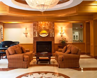 A cozy living room area with two striped armchairs facing each other across a round wooden coffee table. Behind the chairs is a fireplace with a marble surround and a small oval mirror above it. The room features warm wood paneling, wall sconces with soft lighting, a grand chandelier on the ceiling, and a piano in the corner.