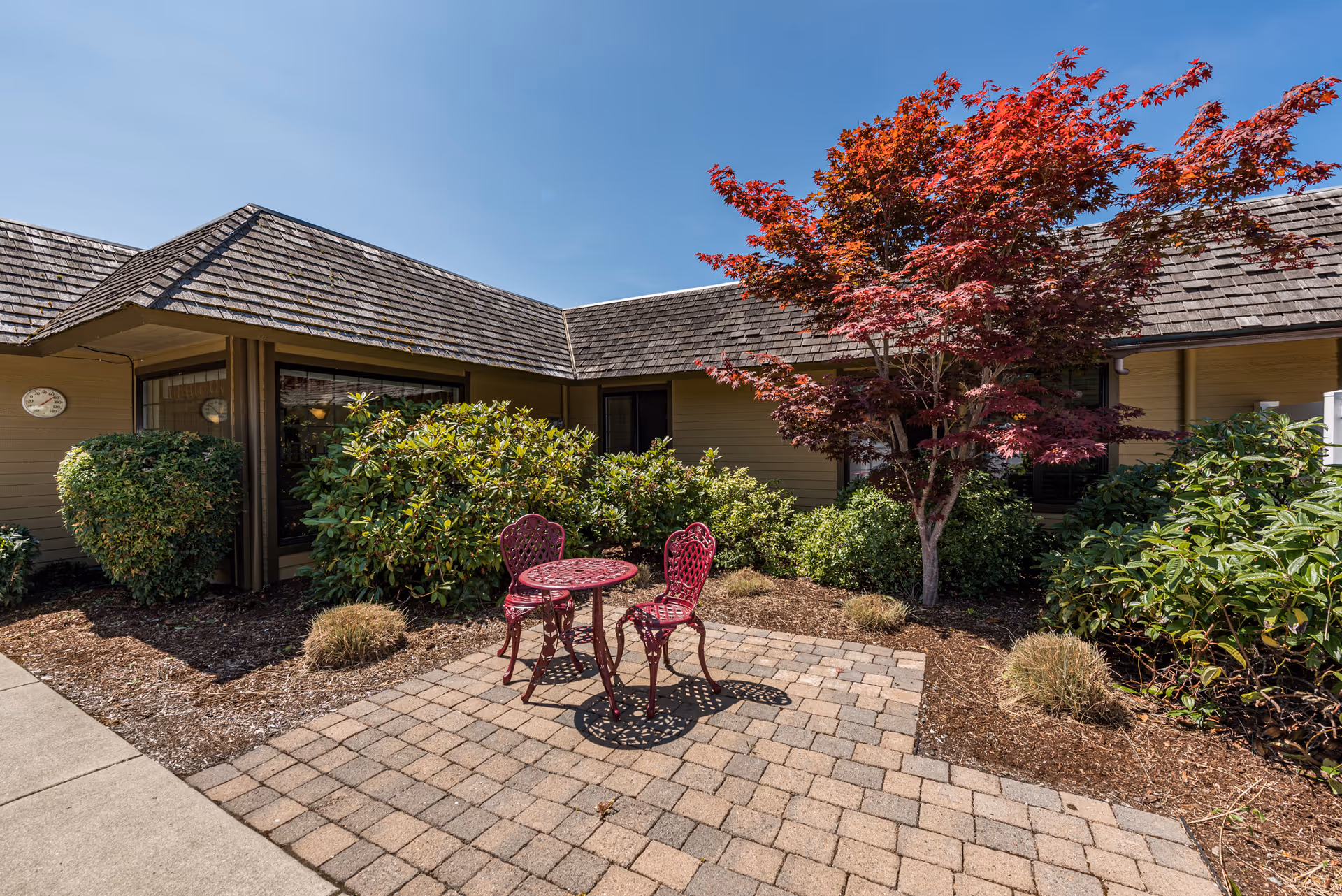 Outdoor patio area with a small red metal table and two matching chairs on a paved stone surface, surrounded by green bushes and a tree with red leaves, adjacent to a building with a shingled roof under a clear blue sky.