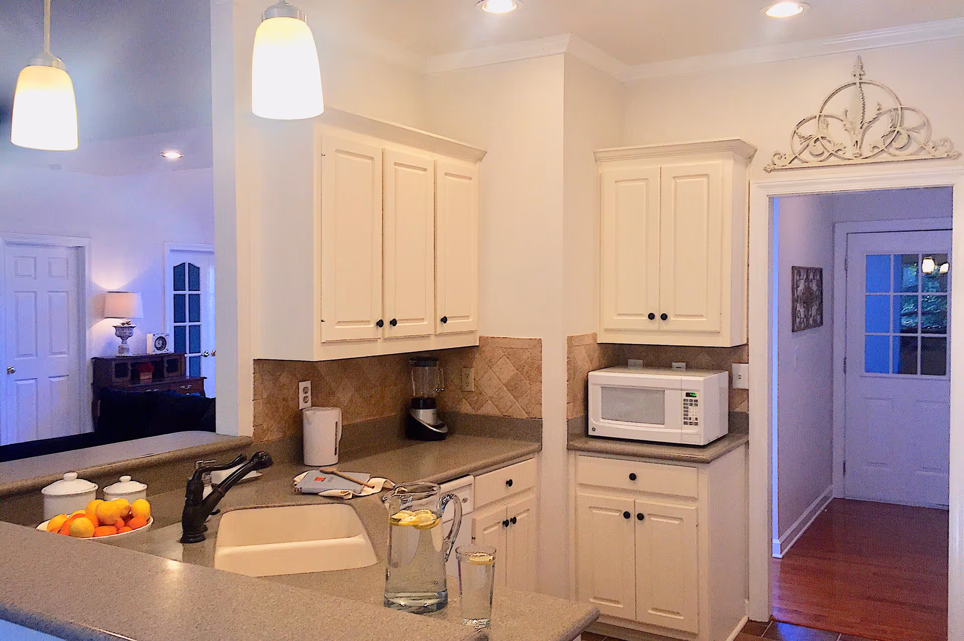 A bright kitchen area with white cabinets, a beige countertop, and a built-in sink. On the counter, there is a pitcher and glass of water with lemon slices, a bowl of oranges, a blender, and a microwave. Two pendant lights hang from the ceiling, and a hallway with a white door is visible in the background.