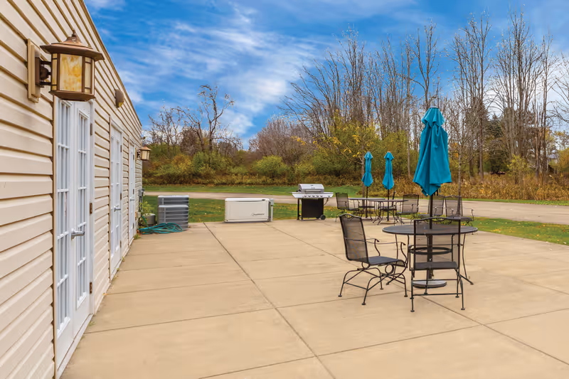 Outdoor patio area with several black metal tables and chairs, each table having a closed turquoise umbrella. The patio is adjacent to a beige building with white doors and wall-mounted lantern-style lights. In the background, there is a grill, some equipment, and a grassy area with trees under a partly cloudy sky.