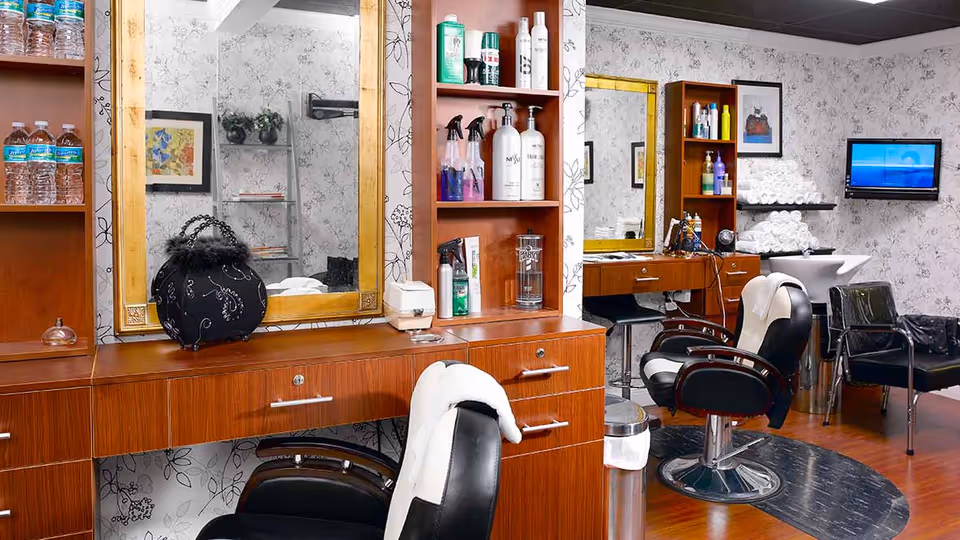 Interior view of a salon area in a senior living facility with wooden cabinetry, large mirrors framed in gold, black salon chairs with white towels draped over them, shelves stocked with hair care products and water bottles, a sink for washing hair, neatly stacked white towels, and a wall-mounted television.