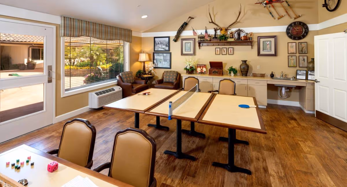 A well-lit activity room with wooden flooring featuring a table tennis setup on two tables, several chairs, a window with blinds, a door leading outside, and a wall decorated with framed pictures, a clock, and various decorative items including a mounted deer head and a bicycle.