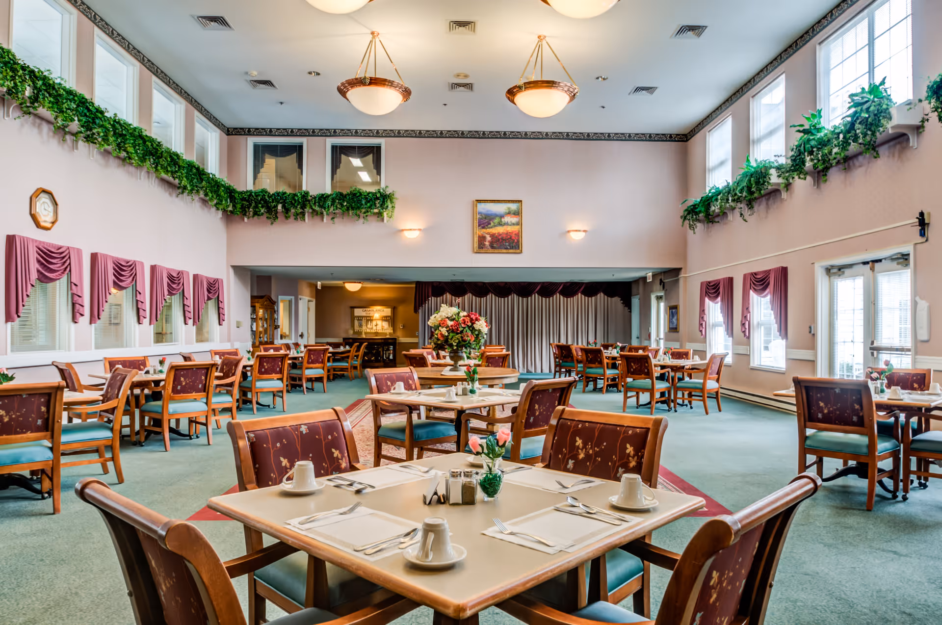 A spacious dining room with multiple tables and chairs arranged neatly. Each table is set with white cups, napkins, and cutlery. The room has large windows with purple drapes and green plants hanging along the upper walls. There are two large ceiling lights and a floral centerpiece on a table in the middle of the room. The walls are light-colored, and there is a framed painting on the far wall.