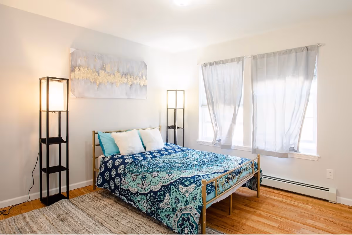 A bright bedroom with a bed featuring a blue and turquoise patterned bedspread and two white pillows. The room has two tall black floor lamps on either side of the bed, light-colored walls, two windows with sheer white curtains, and a light-colored rug on a wooden floor.