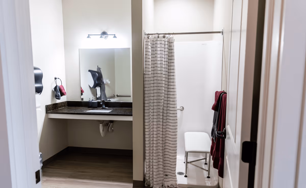 A bathroom with a sink and countertop on the left side, a large mirror above the sink, and a shower area with a striped shower curtain and a white shower chair inside. There are towels hanging on a rack on the right wall.