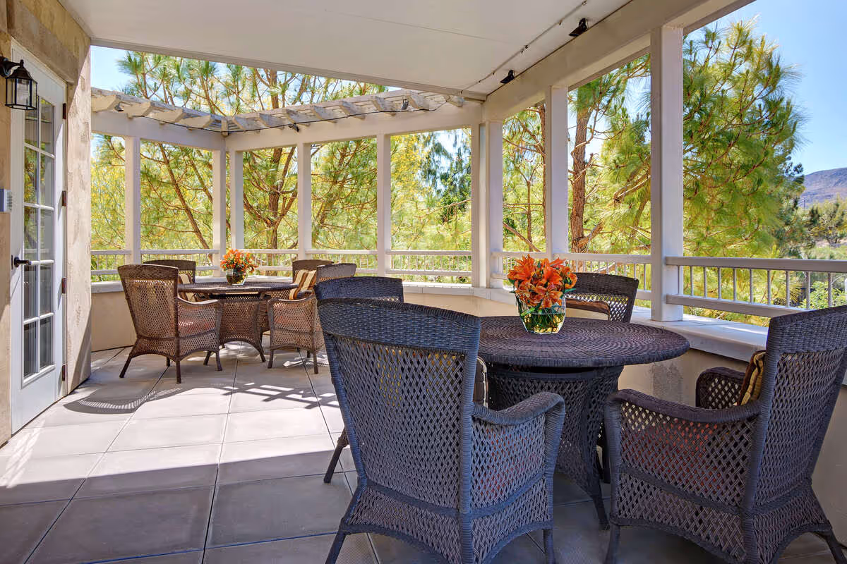 Outdoor covered patio area with two round tables, each surrounded by four wicker chairs. Each table has a vase with orange flowers. The patio is surrounded by white railings and columns, with trees and a clear blue sky visible in the background.