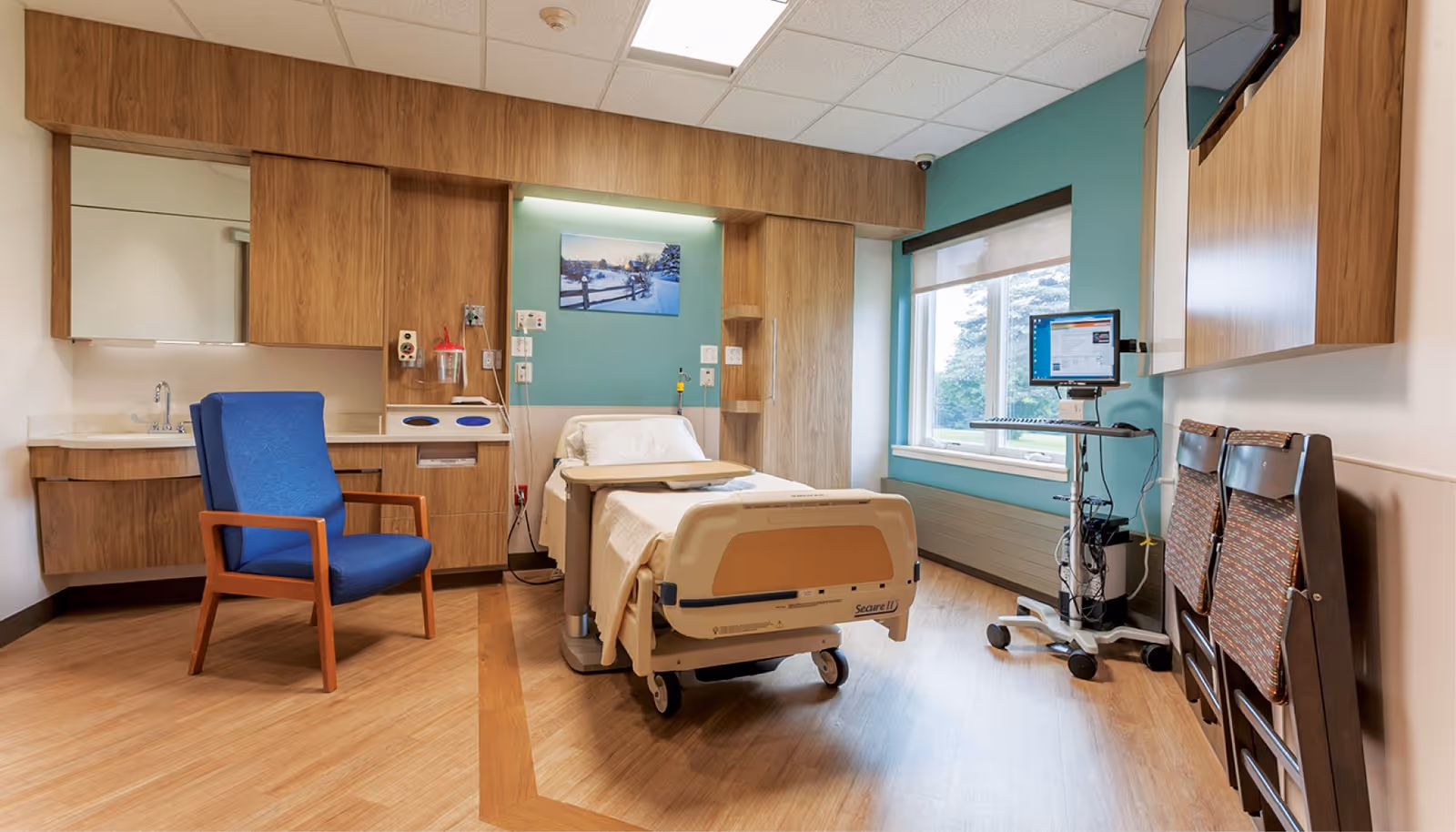 A clean and well-lit patient room with a hospital bed, a blue armchair, wooden cabinets, a computer workstation on wheels, and a window with a view of trees outside. The walls are painted in a soft teal and white color scheme, and there is a framed picture above the bed.