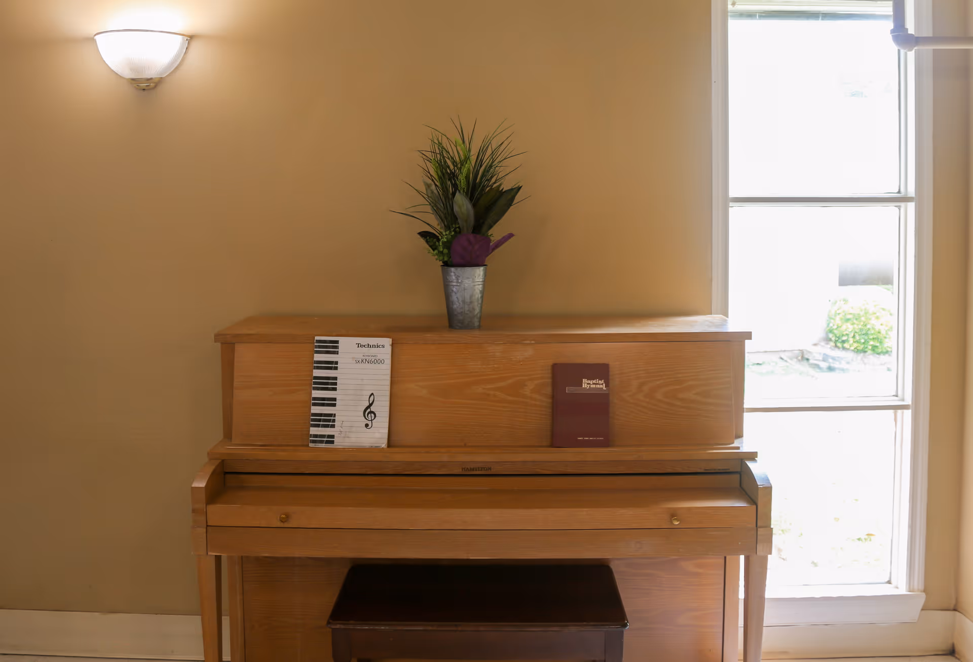 A wooden upright piano against a beige wall with a small potted plant on top and two books resting on the piano's music stand. A wall-mounted light fixture is above the piano, and a large window to the right lets in natural light.