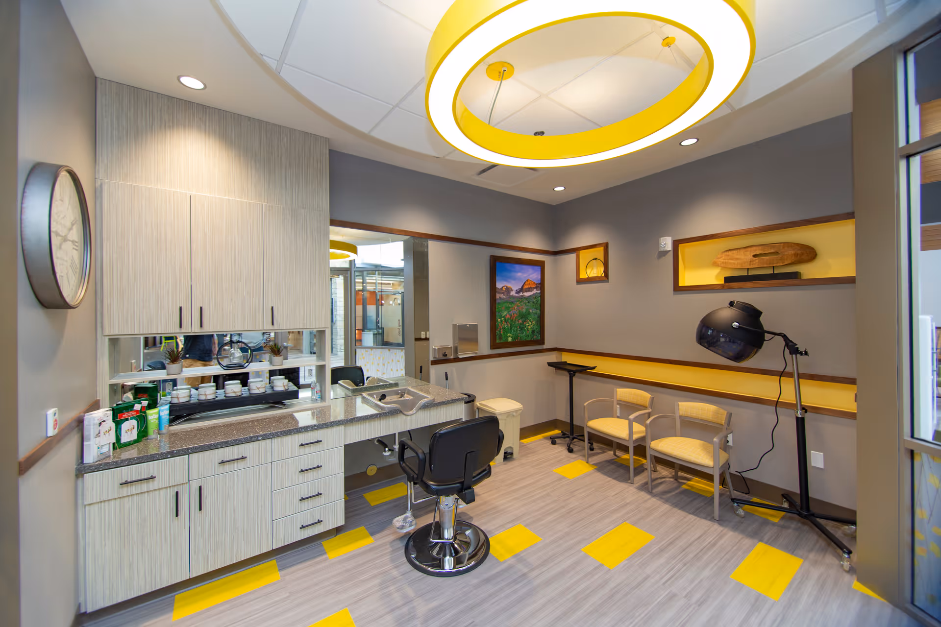 Bright, modern interior room with a salon styling chair and hood dryer, countertop cabinets, and a small waiting area with chairs.