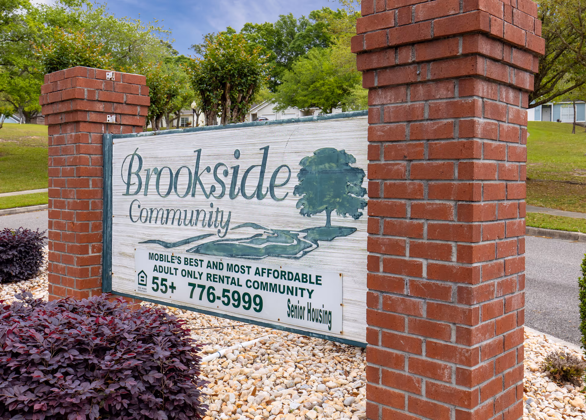 A wooden sign for Brookside Community mounted between two red brick pillars, surrounded by landscaping with purple bushes and white rocks. The sign advertises it as Mobile's best and most affordable adult only rental community for ages 55 and older, with a phone number and mention of senior housing. Trees and a road are visible in the background.