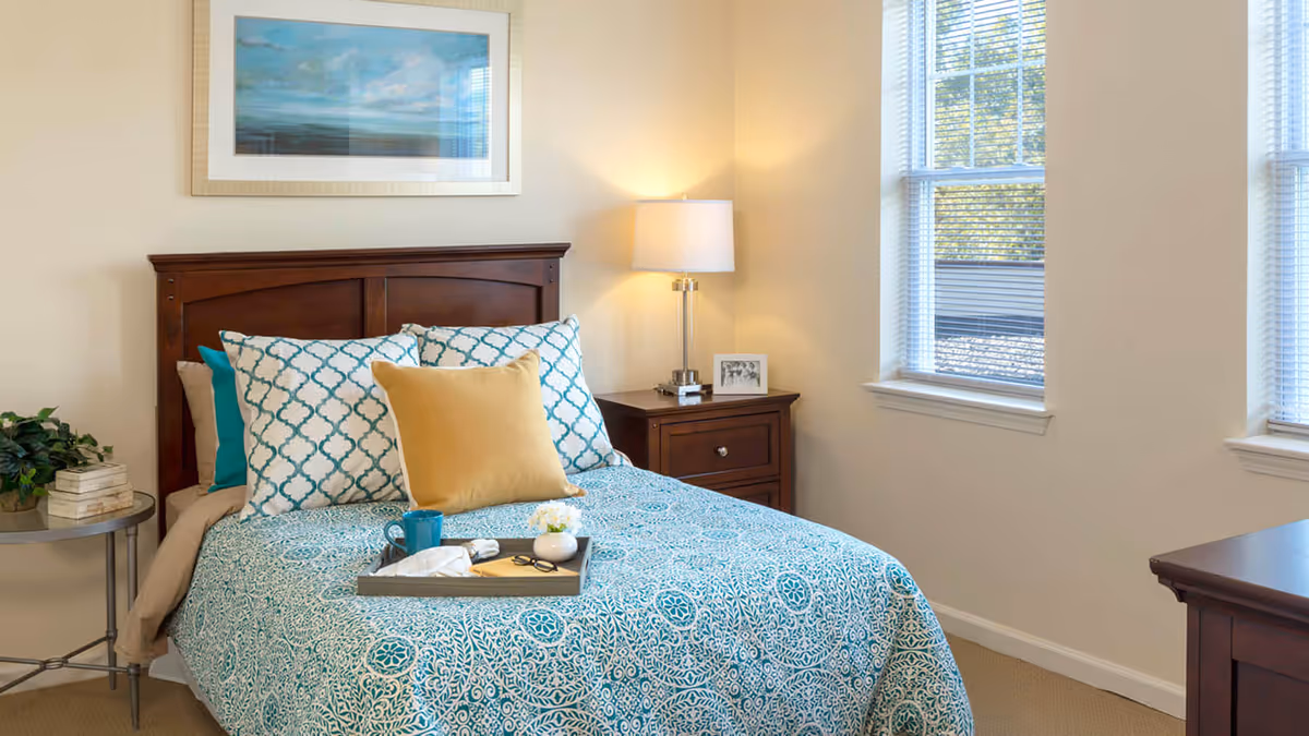 A cozy bedroom with a wooden headboard bed dressed in blue and white patterned bedding, accented with a yellow pillow. A bedside table with a lamp and a framed photo is next to the bed, and a small round table with a plant and books is on the other side. Two windows with blinds allow natural light into the room, and a framed landscape painting hangs above the bed.