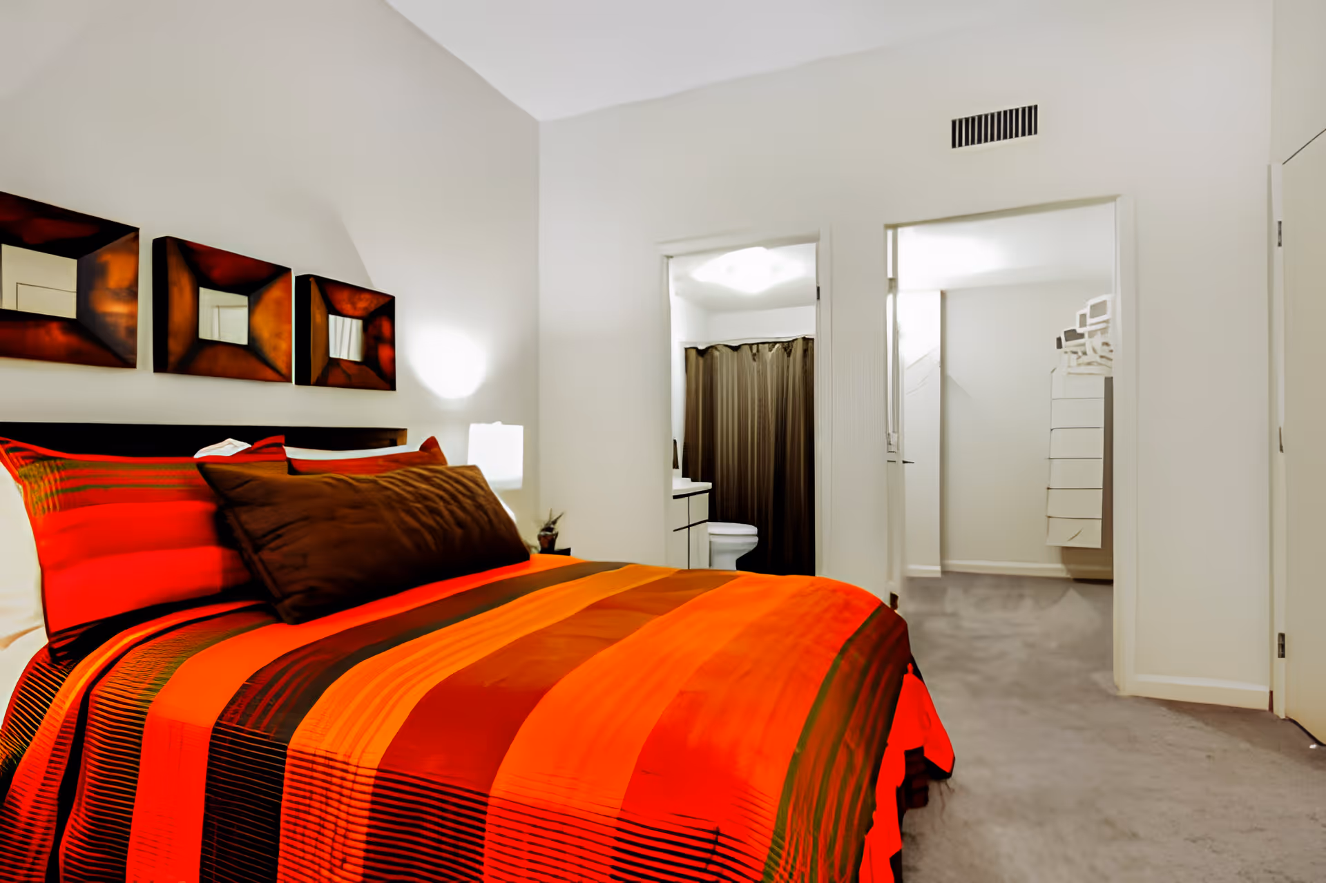 A bedroom with a bed covered in a red, orange, and brown striped bedspread and matching pillows. Above the bed are three square decorative mirrors with dark frames. To the right, there is an open doorway leading to a bathroom with a toilet and a shower curtain. Another open doorway leads to a closet area with shelves and hangers. The walls are white and the floor is carpeted in gray.