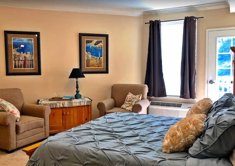 A cozy bedroom in a senior living facility featuring a bed with blue bedding and decorative pillows, two beige armchairs with floral cushions, a wooden side table with a lamp and a tray, two framed paintings on the wall, a window with dark curtains, and a door leading outside.