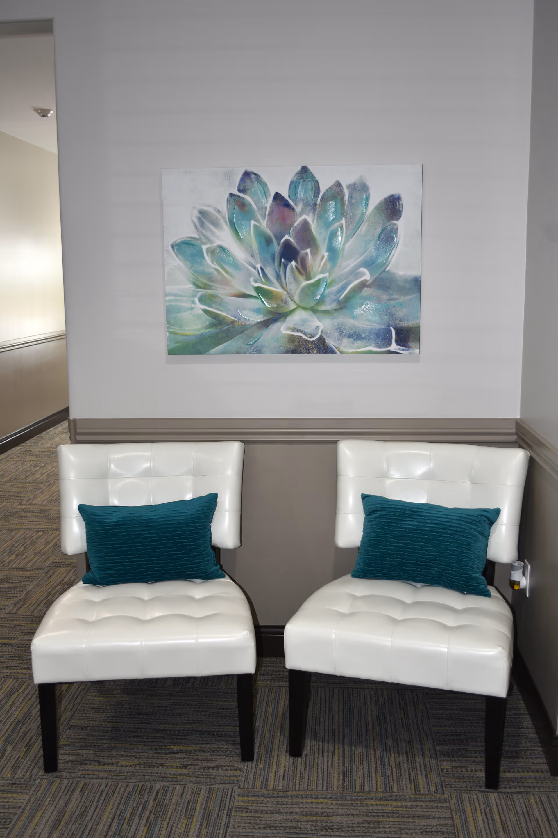Two white cushioned chairs with dark wooden legs, each with a teal pillow, placed side by side against a gray wall. Above the chairs is a large painting of a succulent plant in shades of green, blue, and purple. The floor is carpeted with a patterned design.