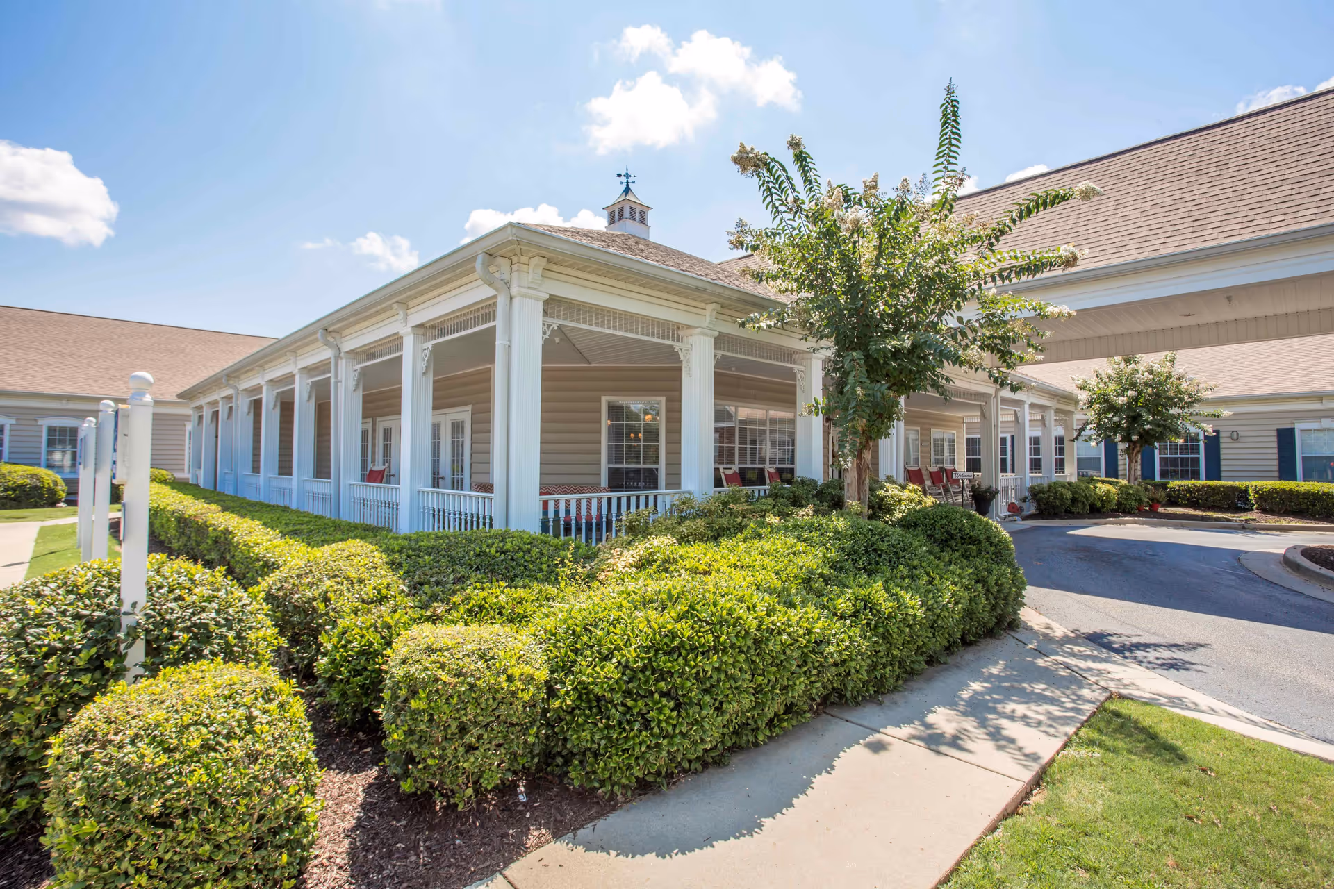 Exterior view of a senior living facility building with beige siding, white columns, and a covered porch area. The porch has several chairs and is surrounded by well-maintained green bushes and small trees. The sky is clear with a few clouds.
