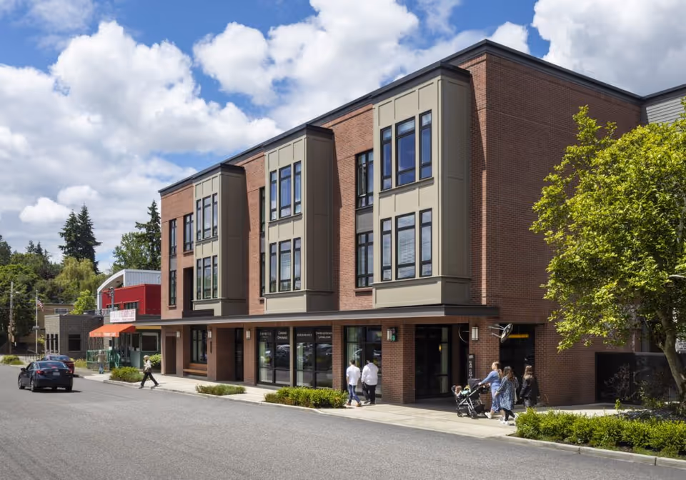 Exterior view of a modern three-story brick building with large windows and a sidewalk in front. Several people are walking on the sidewalk, including a person pushing a stroller. The sky is partly cloudy with blue patches visible.