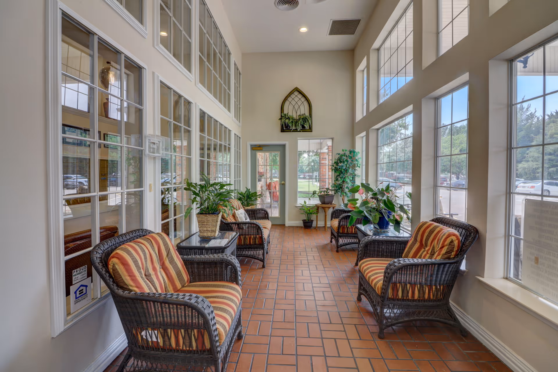 A bright and cozy indoor sitting area with large windows on one side and glass-paneled walls on the other. The space features wicker chairs with striped cushions, small glass-top tables, and several potted plants. The floor is tiled with reddish-brown bricks, and there is a door at the end of the room leading outside.