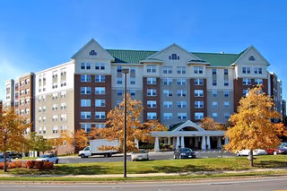 Exterior view of a large multi-story senior living facility building with a green roof, multiple windows, and a covered entrance. There are several cars parked in front and trees with autumn foliage surrounding the building.