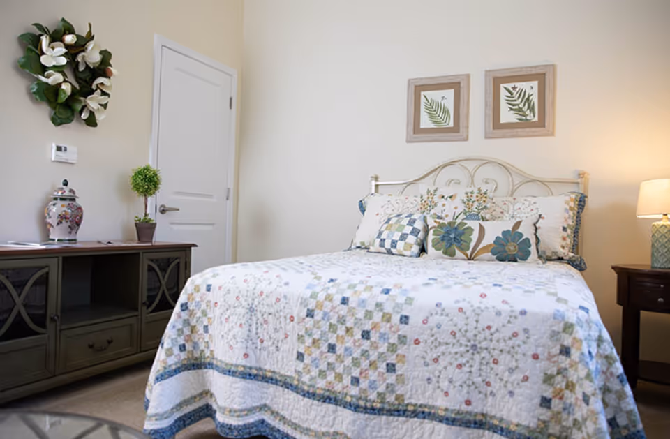 A cozy bedroom with a bed covered in a floral and patchwork quilt, decorated with multiple pillows featuring floral and checkered patterns. Above the bed are two framed botanical prints. To the left is a wooden cabinet with a decorative vase and a small potted plant, and a white door is visible in the background. A table lamp on a nightstand is on the right side of the bed.