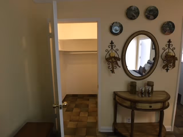 Interior view of a room with an open door leading to a closet with tiled flooring. On the right wall, there is an oval mirror surrounded by decorative plates and candle holders, above a wooden console table with decorative items on it.