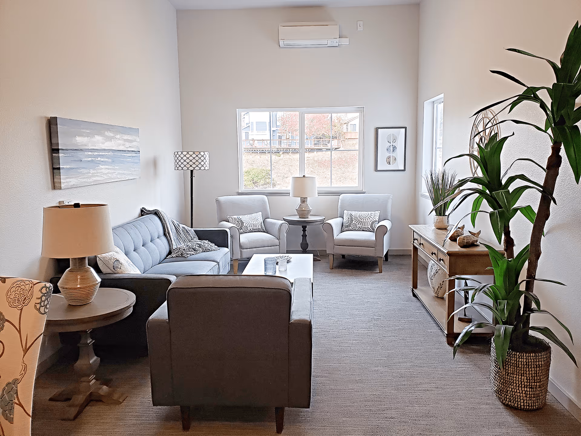 A bright and cozy living room with a gray sofa, two gray armchairs, a gray armchair facing the sofa, a wooden coffee table, and two side tables with lamps. There is a large window letting in natural light, a painting on the wall, and a tall green plant in a woven basket on the right side. A wooden console table with decorative items is against the right wall.