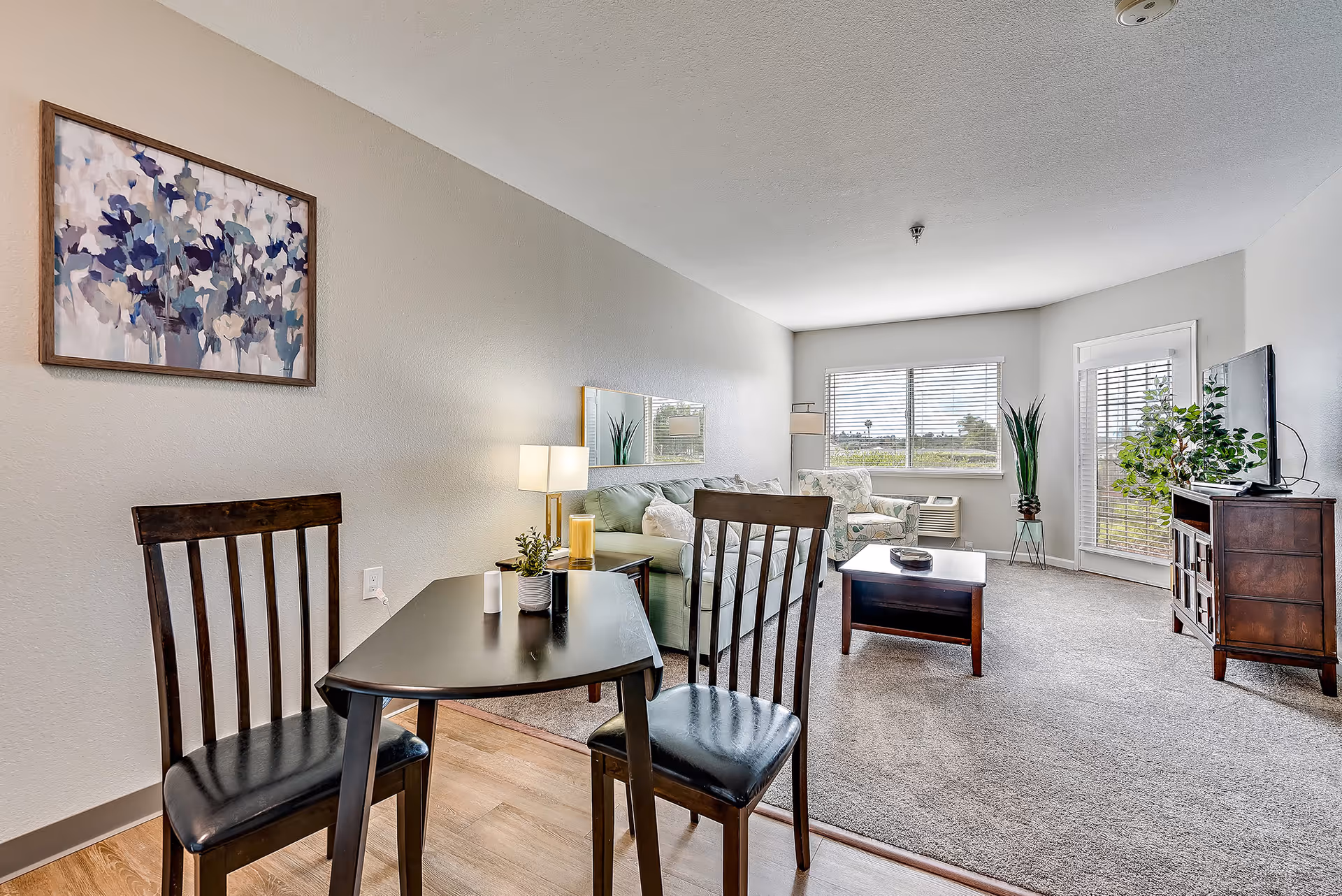 A bright and cozy living room area in a senior living facility featuring a small black dining table with two wooden chairs in the foreground, a green sofa with cushions, a patterned armchair near a window, a wooden coffee table, a TV on a wooden stand, and decorative plants. The room has light-colored walls, carpeted flooring, and a large window letting in natural light.