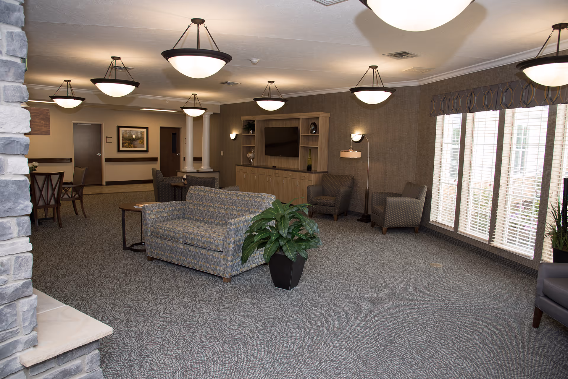 Spacious furnished common lounge with sofas, armchairs, a TV cabinet, large windows, and hanging pendant lights.
