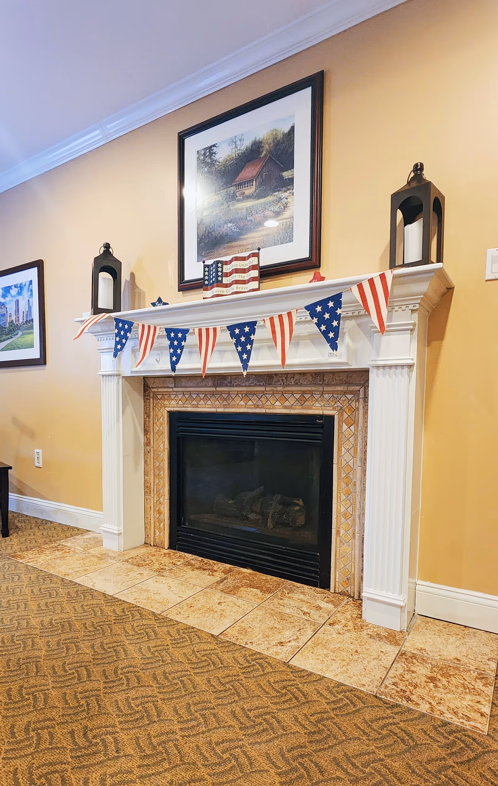 A cozy indoor fireplace with a white mantel decorated with a patriotic red, white, and blue star-spangled banner. On the mantel, there are two black lanterns with white candles inside and a small American flag decoration. Above the fireplace hangs a framed painting of a rustic house surrounded by trees and flowers. The walls are painted a warm yellow, and the floor has a combination of patterned carpet and tile.