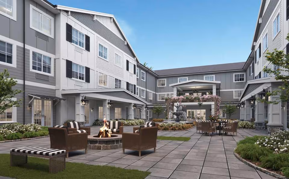 Outdoor courtyard of The Inn at University Village Assisted Living Community featuring a paved patio with a fire pit surrounded by wicker chairs with striped cushions, a dining table with chairs, a central fountain adorned with flowers, and a three-story building with numerous windows and gray siding under a clear blue sky.