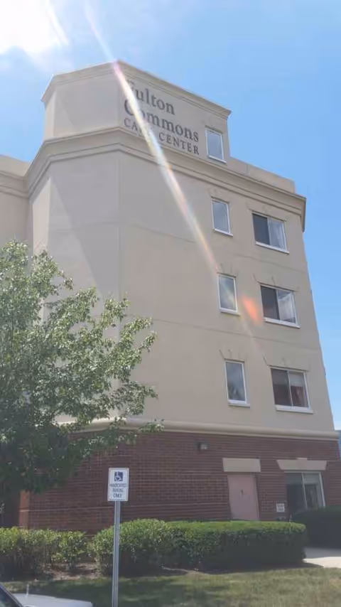 Front exterior of Fulton Commons Care Center building with sign and windows under blue sky.