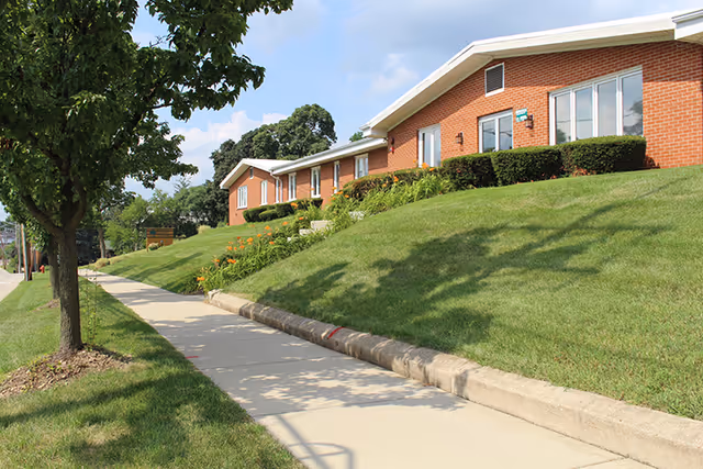 Sidewalk running alongside a grassy slope with a row of single-story brick buildings with windows and bushes under a partly cloudy sky.