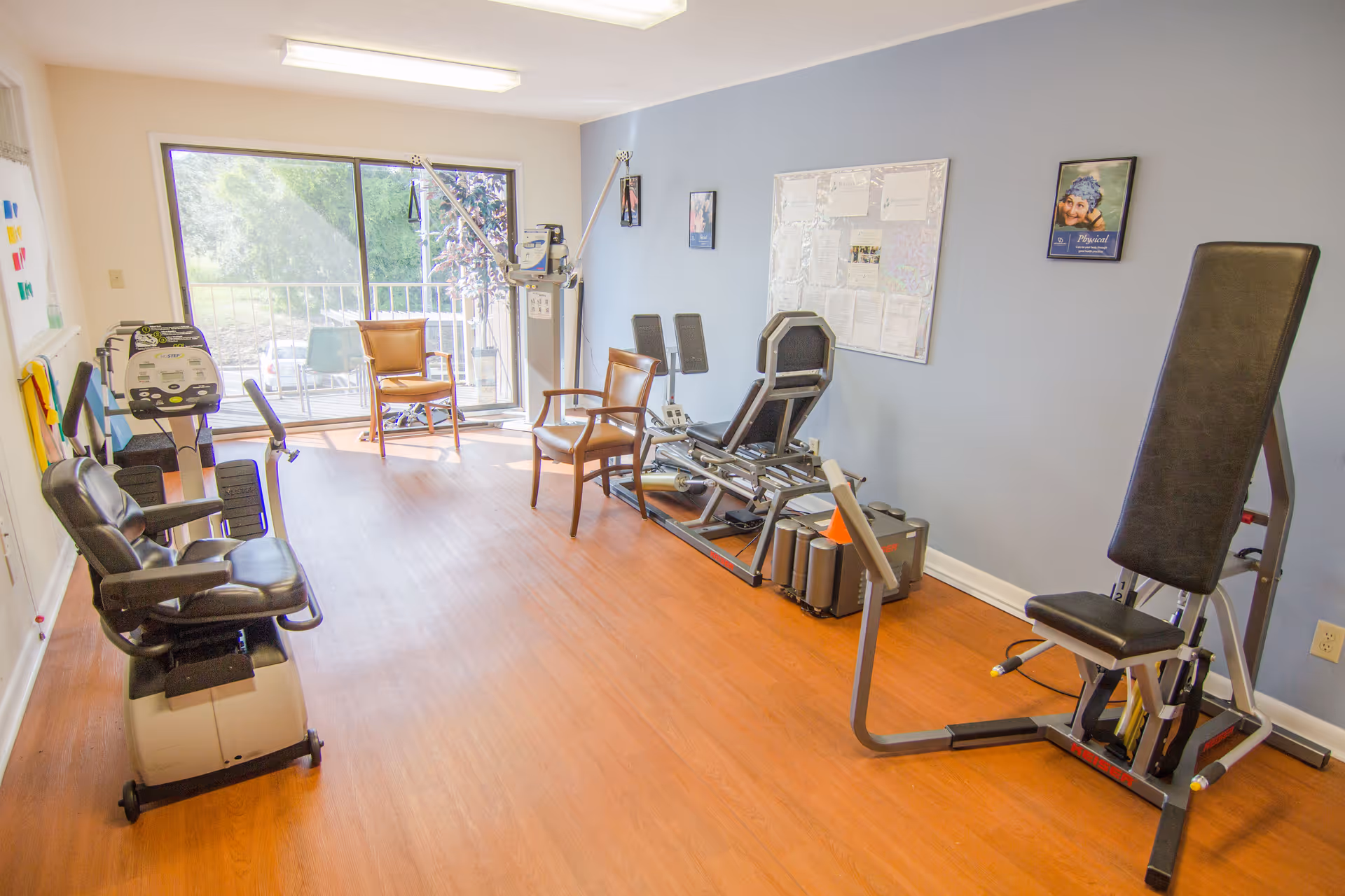 A small exercise room with various fitness equipment including a recumbent bike, leg press machine, and other workout machines. The room has wooden flooring, light blue walls with framed pictures and a bulletin board, and a large sliding glass door letting in natural light and showing outdoor greenery.
