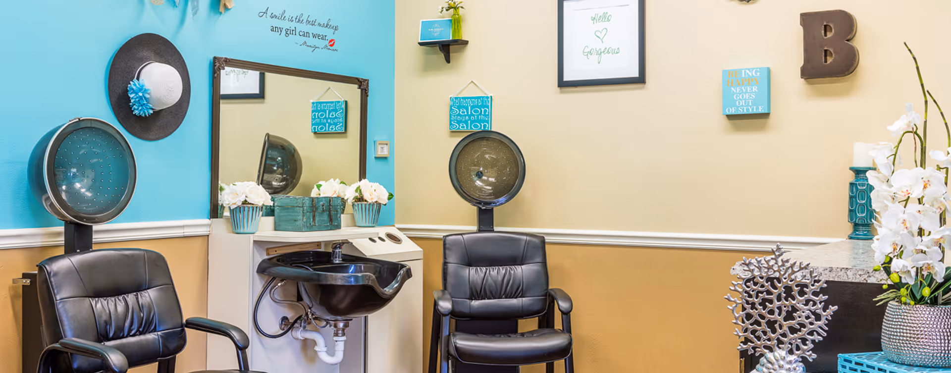 Interior of a salon area in a senior living facility with two black salon chairs facing hair drying stations. The walls are decorated with hats, framed quotes, and decorative items. There is a large mirror above a sink with flowers and a small green box on the counter.