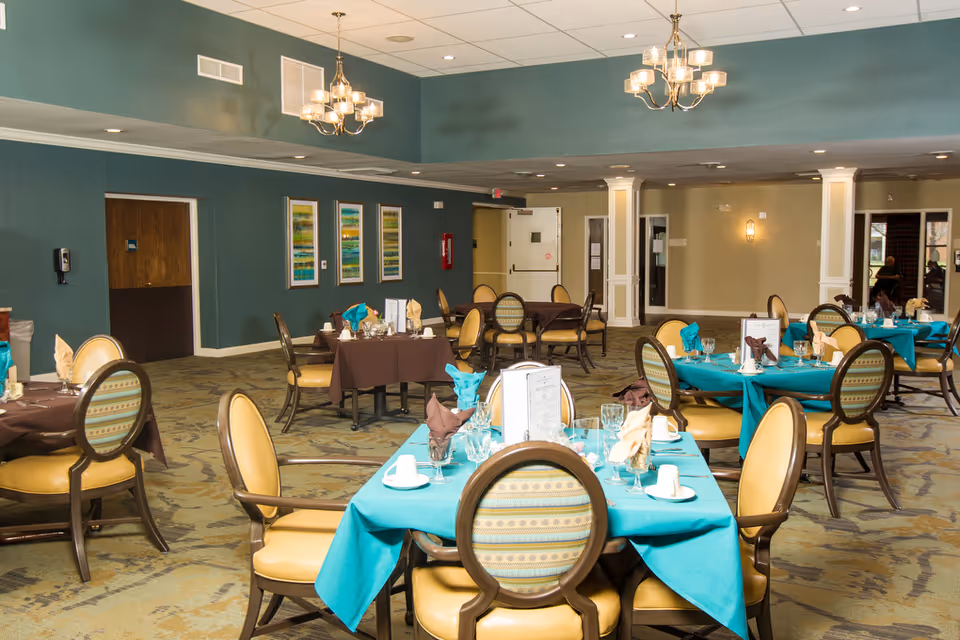 Spacious dining room with round tables set with teal and brown tablecloths, upholstered chairs, and chandeliers.