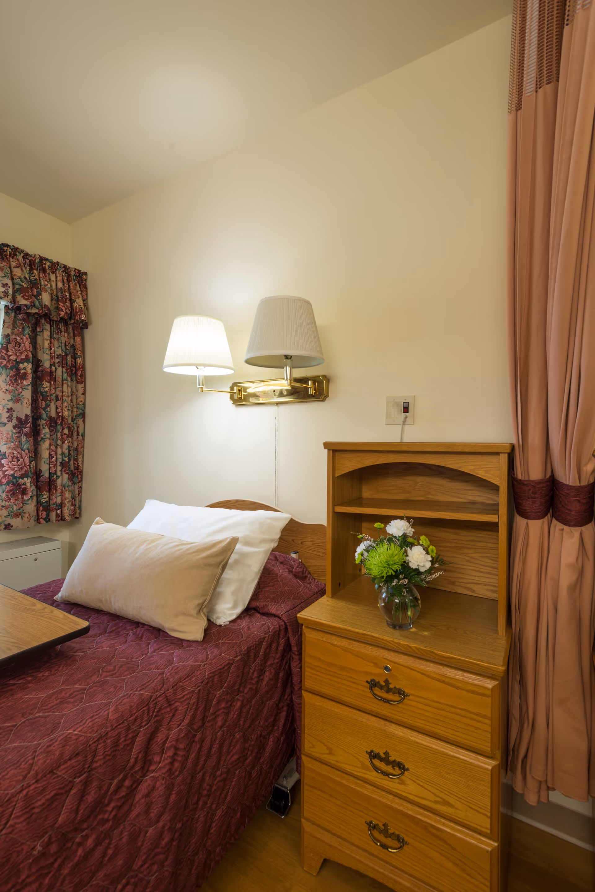 A small bedroom in a skilled nursing facility with a single bed covered in a maroon quilt and two pillows. A wooden nightstand with three drawers and a small shelf holds a glass vase with green and white flowers. Above the bed is a wall-mounted lamp with two white lampshades. Floral curtains cover the window on the left, and peach-colored curtains are tied back on the right.