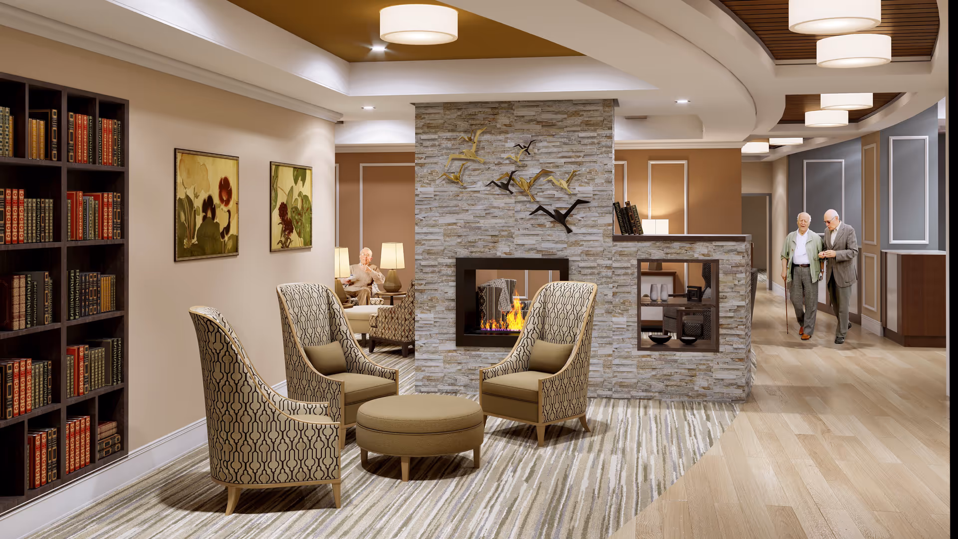 A cozy assisted living facility common area with a stone fireplace in the center, surrounded by three patterned armchairs and a round ottoman. There is a bookshelf filled with books on the left wall, two framed floral paintings, and warm lighting from ceiling fixtures and table lamps. Two elderly men are walking down a hallway on the right side, and another elderly man is seated in the background near the fireplace.