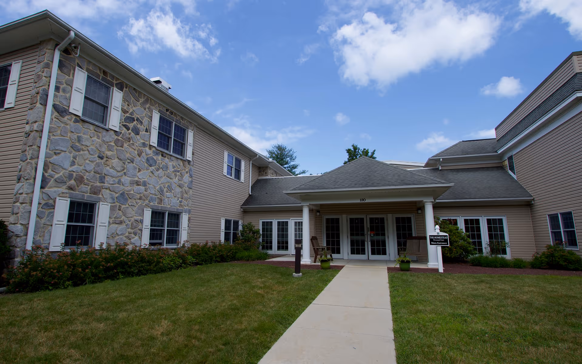 Exterior view of a senior living facility building with stone and beige siding, white window shutters, a covered entrance with columns, a concrete walkway leading to the entrance, and a well-maintained lawn under a partly cloudy sky.