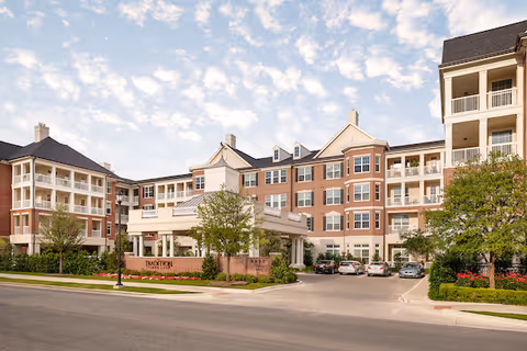 Large multi-story senior living building with a covered entrance, balconies, and landscaped driveway under a partly cloudy sky.