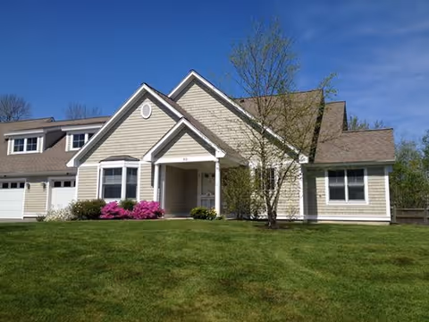 A beige two-story house with white trim and a gabled roof, surrounded by a green lawn and some small trees and bushes, under a clear blue sky.