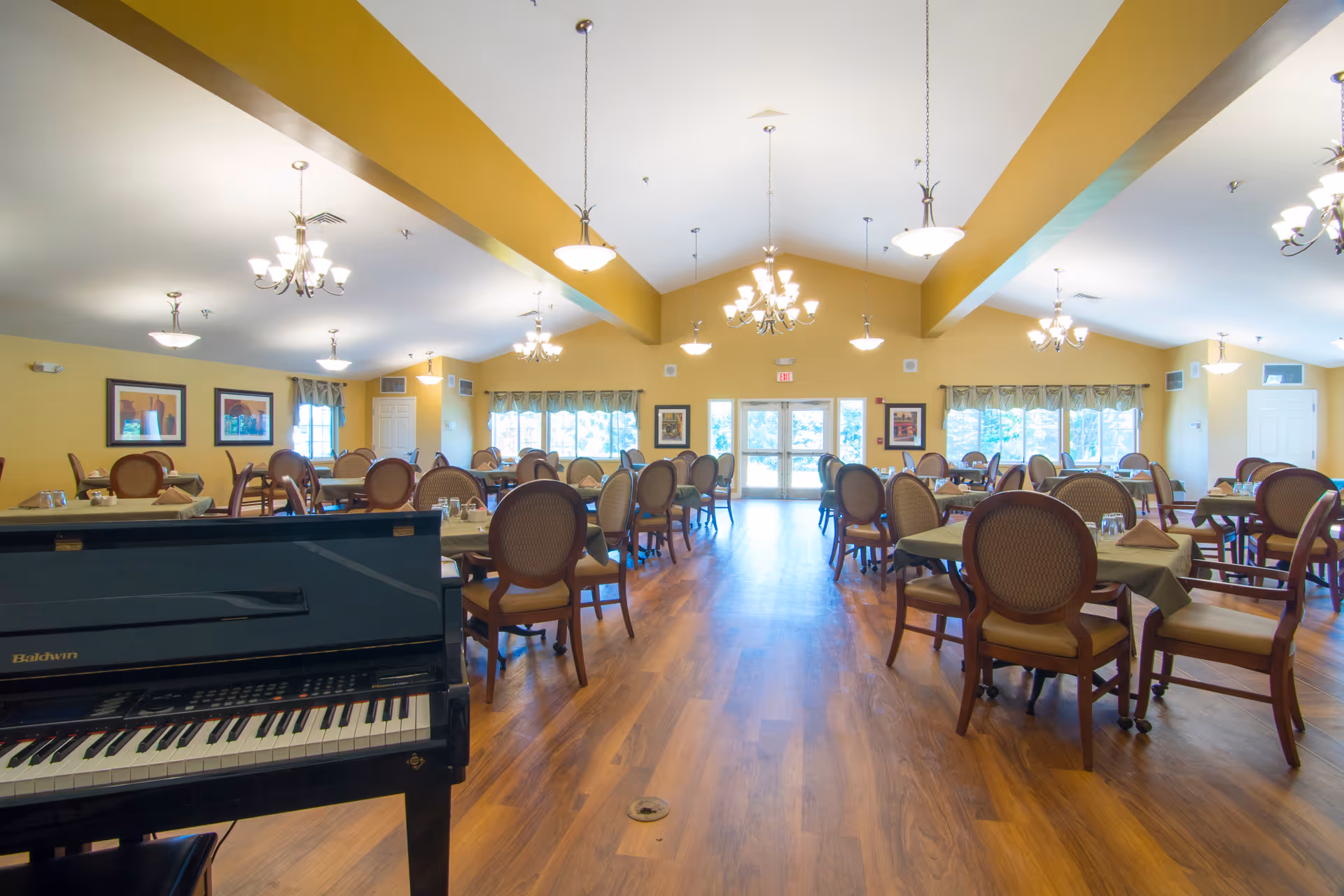 A spacious dining room with multiple tables and chairs arranged neatly. The room has wooden flooring, yellow walls, and several chandeliers hanging from the ceiling. Large windows at the far end let in natural light. A black Baldwin piano is visible in the foreground on the left side.