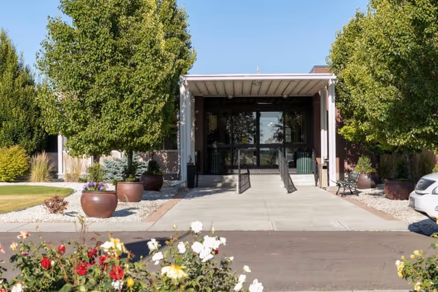 Covered front entrance of a care facility with a wheelchair ramp, potted plants, trees, and flower beds.