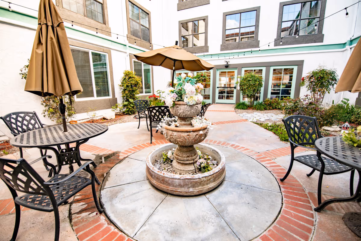 Sunny courtyard with a central tiered fountain surrounded by metal tables, chairs, umbrellas and plants in a senior living community.