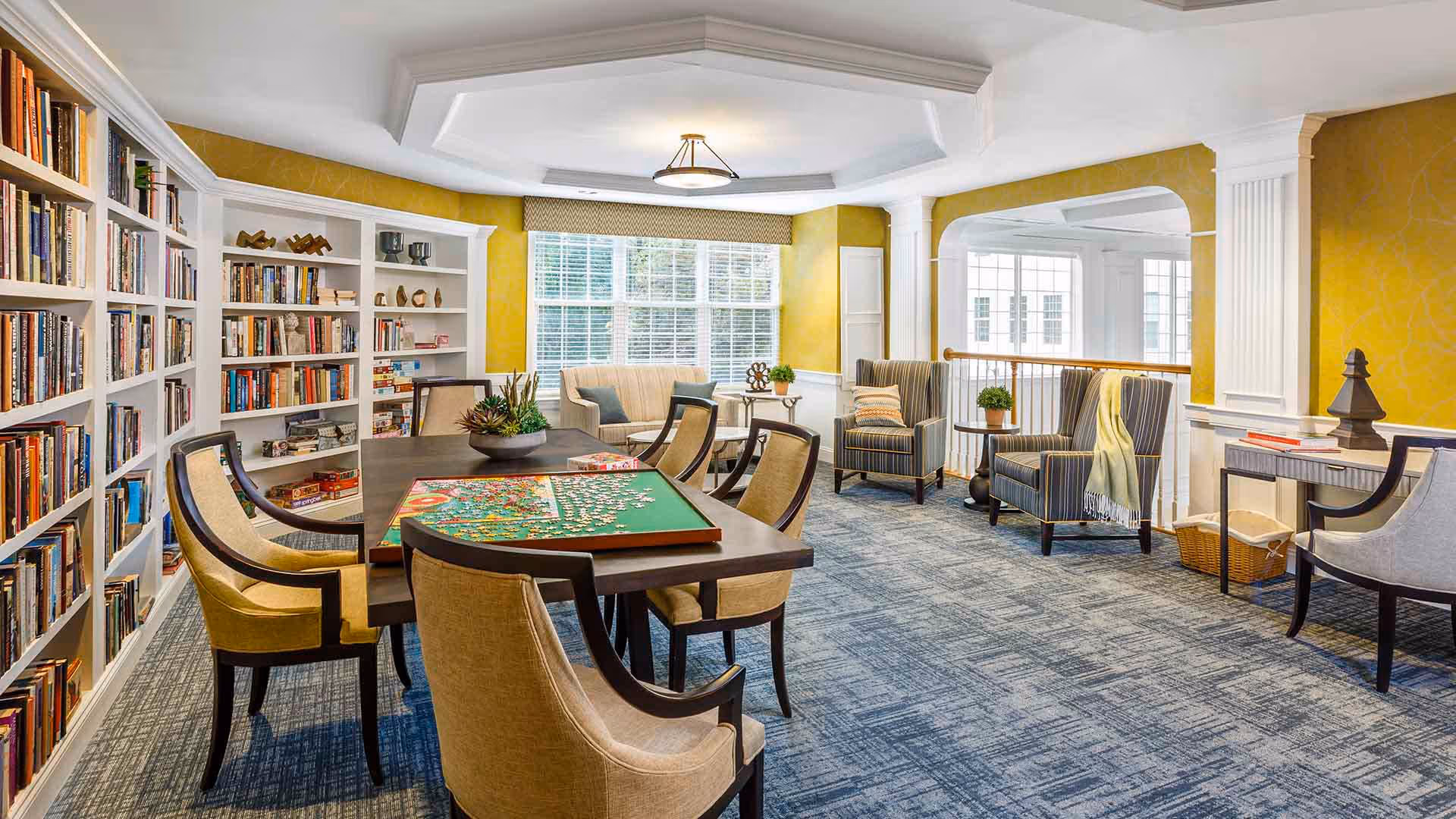 Bright communal lounge with built-in bookshelves, a table with a jigsaw puzzle and surrounding upholstered chairs by large windows.