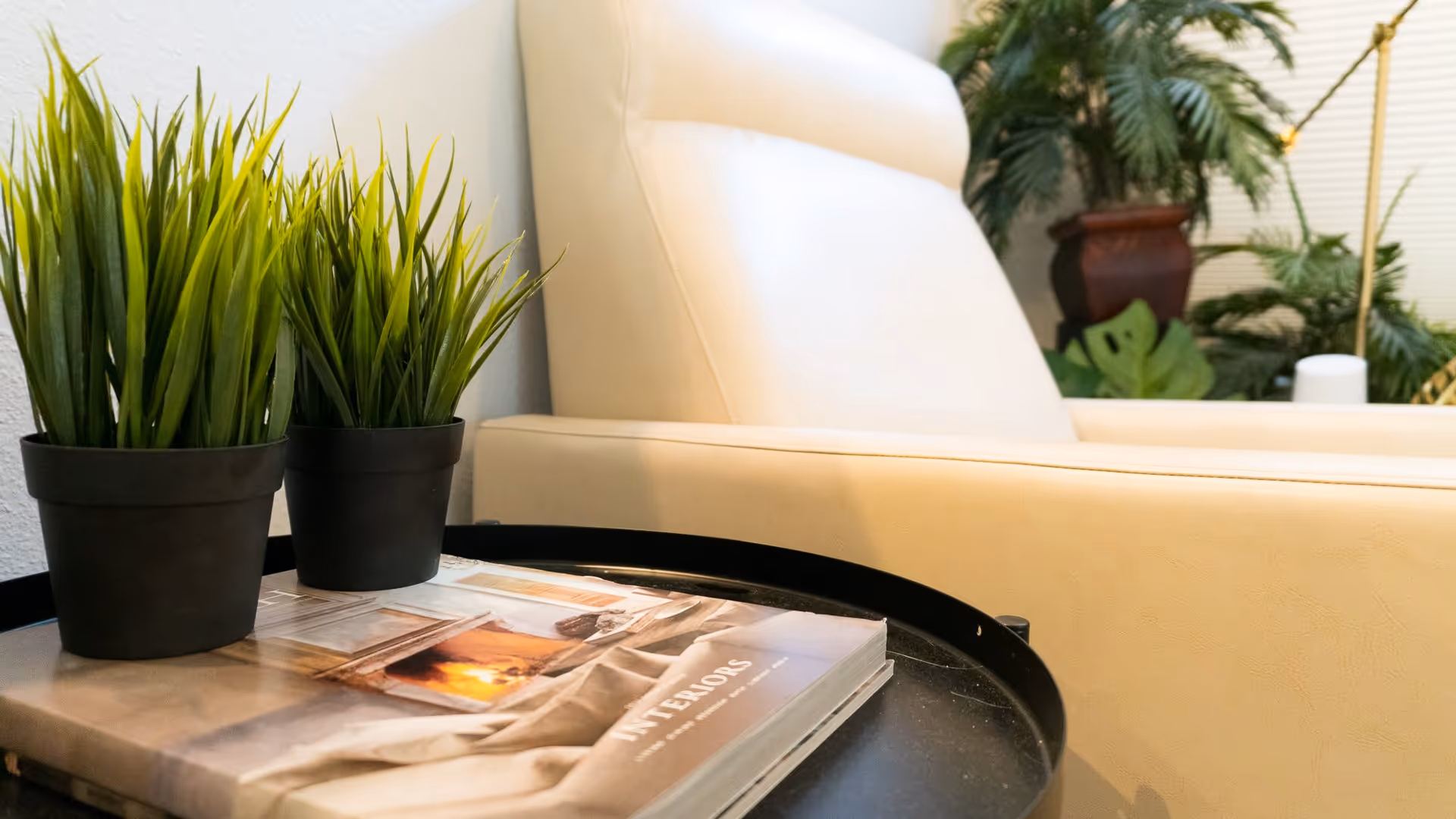 Close-up view of a beige leather chair next to a black round table with two small potted green plants and a book titled 'INTERIORS'. In the background, there are more green plants and a window with blinds.